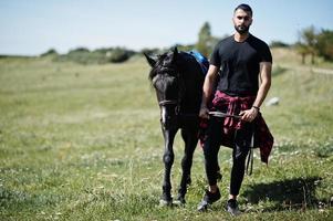 homem árabe de barba alta usa preto com cavalo árabe. foto