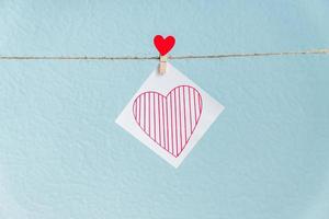 pino de corações de amor do dia dos namorados vermelho pendurado no cordão natural contra um fundo azul. coração desenhado em pedaço de papel. foto