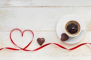 xícara de café americano preto com chocolate banhado a ouro em forma de coração e fita de cetim vermelha em fundo de madeira. vista do topo. foto