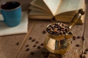grãos de café torrados frescos no bule de café turco tradicional cezve abriu o livro e a xícara na mesa de madeira. foto