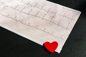 close-up de um eletrocardiograma em papel com coração de madeira vermelho. papel ecg ou ekg em fundo preto. conceito médico e de saúde. foto