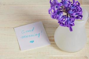 etiqueta de papel de bom dia e flor de jacinto roxo em vaso branco na mesa de madeira foto