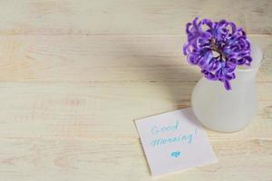 etiqueta de papel de bom dia e flor de jacinto roxo em vaso branco na mesa de madeira foto