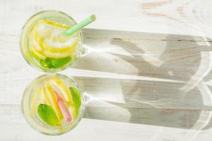 copo de limonada caseira com limões, hortelã e canudos de papel em fundo rústico de madeira. bebida refrescante de verão. sombras duras foto