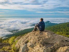 homens asiáticos sentam-se no penhasco com lindo céu do nascer do sol na montanha khao luang no parque nacional de ramkhamhaeng, província de sukhothai tailândia foto