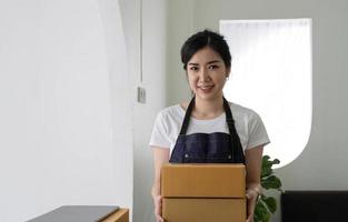 empreendedor de pequenas empresas de inicialização de mulher asiática freelance usando um laptop com caixa alegre mulher asiática sua mão levanta caixa de embalagem de marketing on-line e conceito de ideia de entrega sme foto