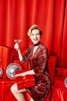 mulher elegante elegante madura com copo de vinho espumante com presente em fundo vermelho. festa, moda, celebração, conceito anti-idade foto