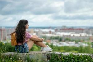 uma garota turista em uma camiseta rosa com uma mochila laranja está sentada e apreciando a vista panorâmica. foto