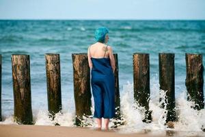 jovem mulher de cabelos azuis com vestido longo azul escuro em pé na praia, olhando para o horizonte do mar