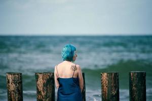 linda mulher de cabelos azuis com vestido longo azul escuro em pé na praia, olhando para o horizonte do mar