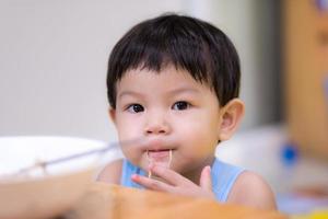 uma criança de 2 anos usa a mão para pegar macarrão instantâneo. menino asiático colocou uma linha de macarrão instantâneo na boca e olhou para a câmera. filho está comendo comida em casa. foto