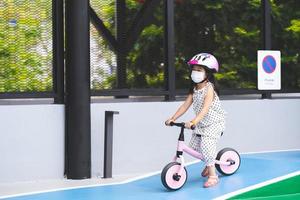 imagem de retrato criança de 5 anos. linda garota asiática usando máscara facial branca está andando de bicicleta de arado rosa pequena colina no campo de treinamento, sinal de trânsito com tailandês e inglês escrevendo sem estacionamento e símbolo. foto