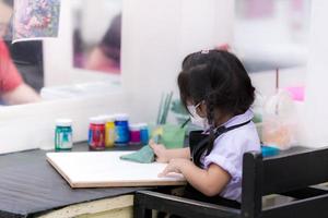 aluno usando um pano para limpar uma tela. criança se preparando para aprender a pintar aquarela por tutor especial que seus pais trouxeram para estudar depois da escola. criança asiática de 3 anos de idade usa máscara facial. foco seletivo foto