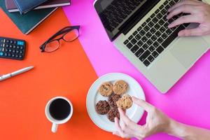 mãos masculinas segurando biscoitos e usando laptop, xícara de café e material de escritório em fundo colorido. foto