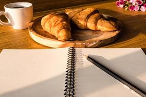 caderno em branco com lápis, croissant e café no fundo da mesa de madeira. foto