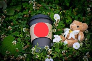 copo de papel pardo para café e ursinho de pelúcia na grama verde. xícara de café com urso de brinquedo na grama. foto