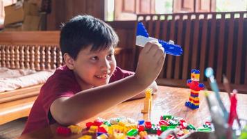 menino asiático está ficando criativo com a montagem de tijolos de plástico coloridos em robôs e aviões em uma mesa de madeira feliz e divertido no conceito de criadores home.kid. foto