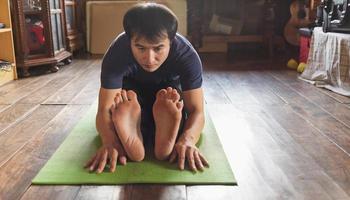 jovem asiático praticando ioga, sentado no exercício de flexão para a frente sentado, pose de paschimottanasana na ioga de esteira verde em casa de madeira. vida saudável foto