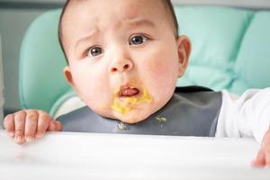 um bebê sujo e surpreso manchado de purê de legumes na mesa de alimentação. introdução de alimentos complementares, não gosto do sabor, comida nova. close-up, retrato foto