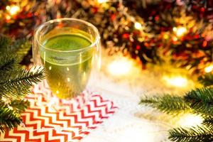 copo de vidro transparente de parede dupla com chá matcha japonês na mesa com decoração de natal. atmosfera de ano novo, guirlanda e enfeites, ramo de abeto, aconchegante, cobertor de malha, bola, guardanapo listrado foto