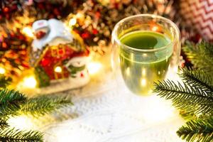 copo de vidro transparente de parede dupla com chá matcha japonês na mesa com decoração de natal. atmosfera de ano novo, guirlanda e enfeites, ramo de abeto, aconchegante, cobertor de malha, bola, guardanapo listrado foto