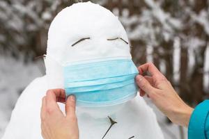 boneco de neve em uma máscara médica - uma nova realidade, proteção contra doenças, infecções, vida na epidemia de covid. as mãos das mulheres colocam uma máscara de boneco de neve. atividades ao ar livre em família de inverno foto