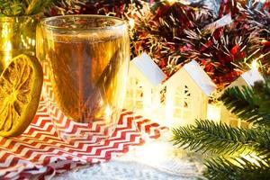 copo de vidro transparente de parede dupla com chá quente e paus de canela em cima da mesa com decoração de natal e casinha. atmosfera de ano novo, fatia de laranja seca, guirlanda, ramo de abeto, aconchegante foto