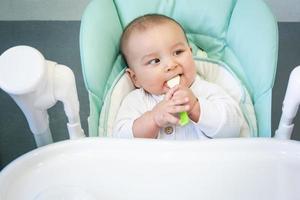 um bebê faminto está roendo uma colher de plástico na mesa em uma cadeira alta. dentição, caprichos, coceira nas gengivas, introdução de alimentos complementares foto