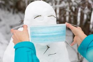 boneco de neve em uma máscara médica - uma nova realidade, proteção contra doenças, infecções, vida na epidemia de covid. as mãos das mulheres colocam uma máscara de boneco de neve. atividades ao ar livre em família de inverno foto