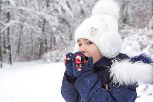 uma garotinha no frio aquece as mãos em luvas - respira ar quente da boca nas mãos. inverno, passear o bebê ao ar livre, na neve. agasalhos, gorro de malha, pompom de pelo e capuz foto