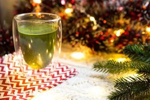 copo de vidro transparente de parede dupla com chá matcha japonês na mesa com decoração de natal. atmosfera de ano novo, guirlanda e enfeites, ramo de abeto, aconchegante, cobertor de malha, bola, guardanapo listrado foto