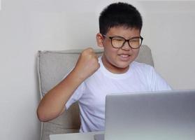 o menino sentado no sofá está assistindo laptop e faz o gesto do vencedor. conceito de educação, aprendizagem e tecnologia. foto