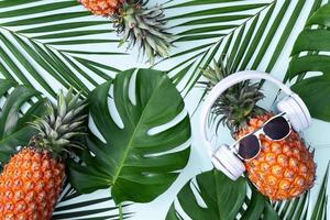abacaxi engraçado usando fone de ouvido branco, conceito de ouvir música, isolado em fundo azul com folhas de palmeira tropicais, vista superior, design plano leigo.