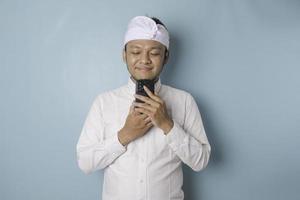 retrato de um jovem balinês pensativo vestindo udeng ou bandana tradicional e camisa branca, olhando de lado enquanto segura o smartphone isolado sobre fundo azul foto