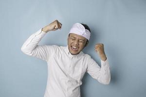 um jovem balinês com uma expressão feliz e bem sucedida usando udeng ou bandana tradicional e camisa branca isolada por fundo azul foto