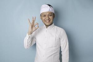 animado homem balinês vestindo udeng ou headband tradicional e camisa branca, dando um gesto de mão ok isolado por um fundo azul foto