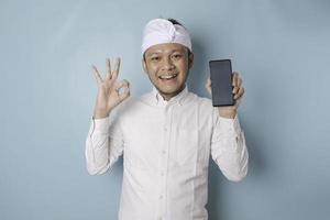 animado homem balinês vestindo udeng ou headband tradicional e camisa branca dá ok gesto de aprovação enquanto segura o smartphone, isolado por fundo azul foto