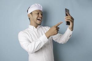 um retrato de um homem balinês feliz está sorrindo e segurando seu smartphone usando udeng ou bandana tradicional e camisa branca isolada por um fundo azul foto