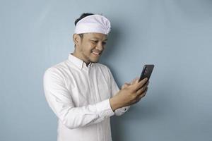 um retrato de um homem balinês feliz está sorrindo e segurando seu smartphone usando udeng ou bandana tradicional e camisa branca isolada por um fundo azul foto