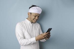 um retrato de um homem balinês feliz está sorrindo e segurando seu smartphone usando udeng ou bandana tradicional e camisa branca isolada por um fundo azul foto