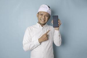 animado homem balinês vestindo tradicional headband ou udeng e camisa branca apontando para o espaço de cópia em seu smartphone, isolado por fundo azul foto