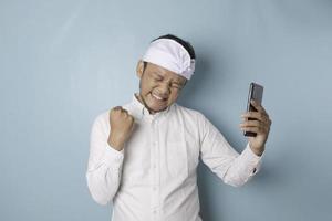 um jovem balinês com uma expressão feliz e bem sucedida usando udeng ou bandana tradicional e camisa branca segurando smartphone isolado por fundo azul foto