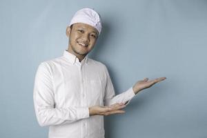 animado homem balinês vestindo udeng ou headband tradicional e camisa branca apontando para o espaço de cópia ao lado dele, isolado por fundo azul foto