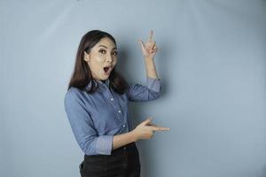 mulher asiática chocada vestindo camisa azul apontando para o espaço de cópia ao lado dela, isolado por fundo azul foto