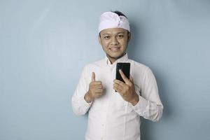 animado homem balinês vestindo udeng ou headband tradicional e camisa branca dá polegares para cima gesto de aprovação enquanto segura o smartphone, isolado por fundo azul foto