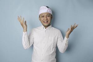 animado homem balinês vestindo udeng ou headband tradicional e camisa branca apontando para o espaço de cópia de cabeça para ele, isolado por fundo azul foto