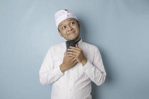 retrato de um jovem balinês pensativo vestindo udeng ou bandana tradicional e camisa branca, olhando de lado enquanto segura o smartphone isolado sobre fundo azul foto