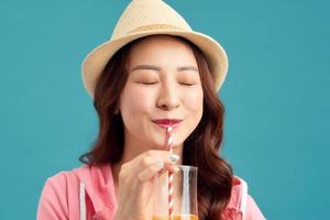 alegre mulher asiática bebendo suco de frutas e usando chapéu de palha, camiseta branca, jeans curtos, jaqueta rosa sobre fundo azul. foto