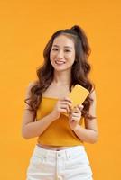 foto de uma linda mulher asiática sorridente segurando o telefone móvel em pé sobre fundo amarelo.