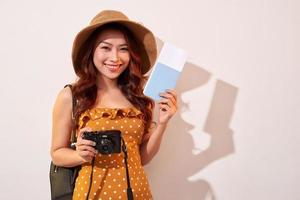 retrato de uma jovem feliz no chapéu segurando a câmera e mostrando o passaporte em pé isolado sobre fundo bege foto